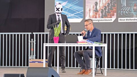 Fabulant und Lehrer auf der Hessentagsbühne Ein Mann mit Hasenmaske, stehend auf der Bühne des Hessentags sowie ein Mann, sitzend vor einem Tisch mit einem Laptop sowie Büchern und einer Topfpflanze darauf.